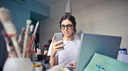 Woman in White T-shirt Holding Smartphone in Front of Laptop